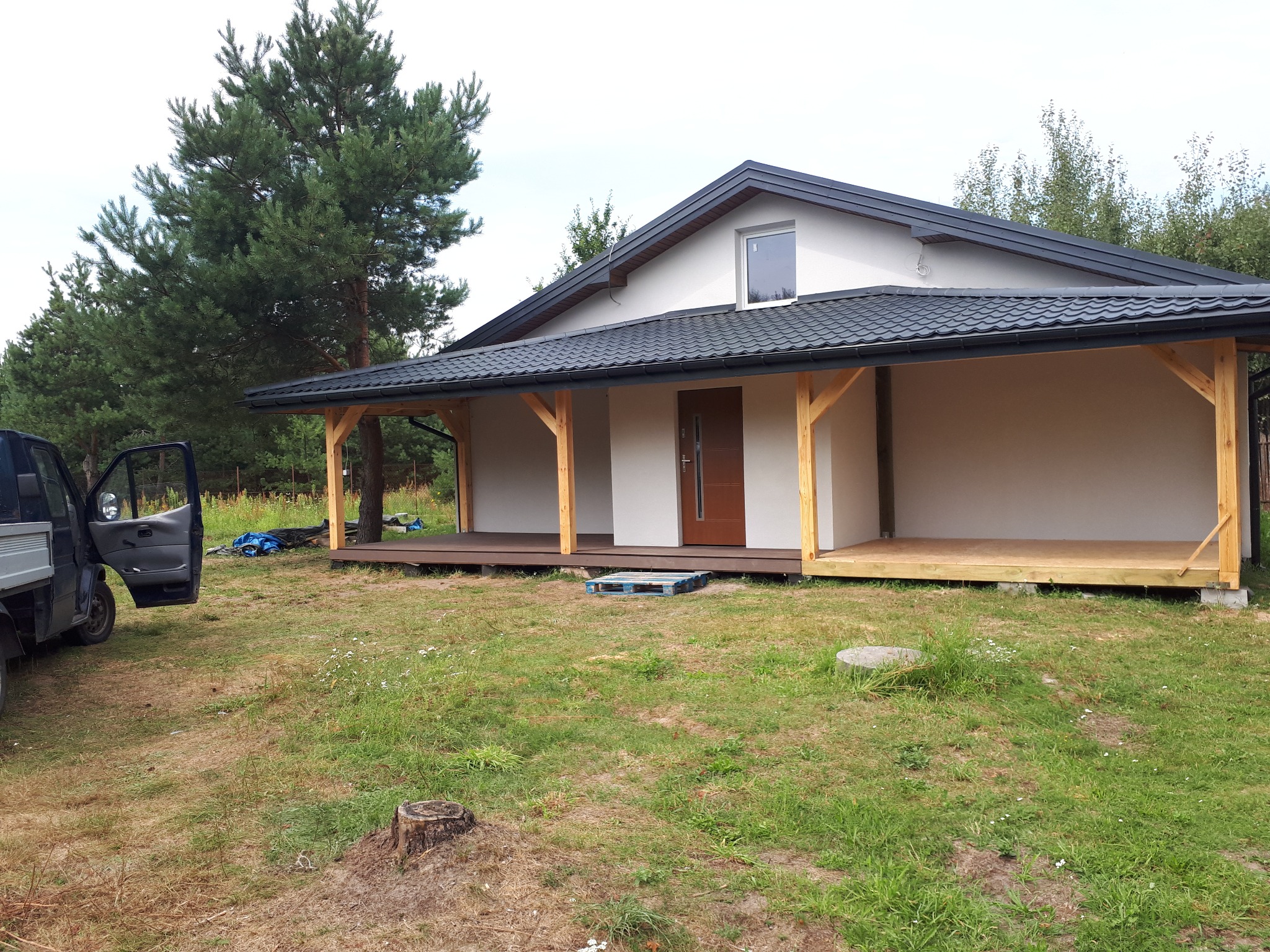 Dom jednorodzinny z nowym, czarnym dachem z blachodachówki, drewnianą konstrukcją werandy i paletą leżącą na trawie przed wejściem.