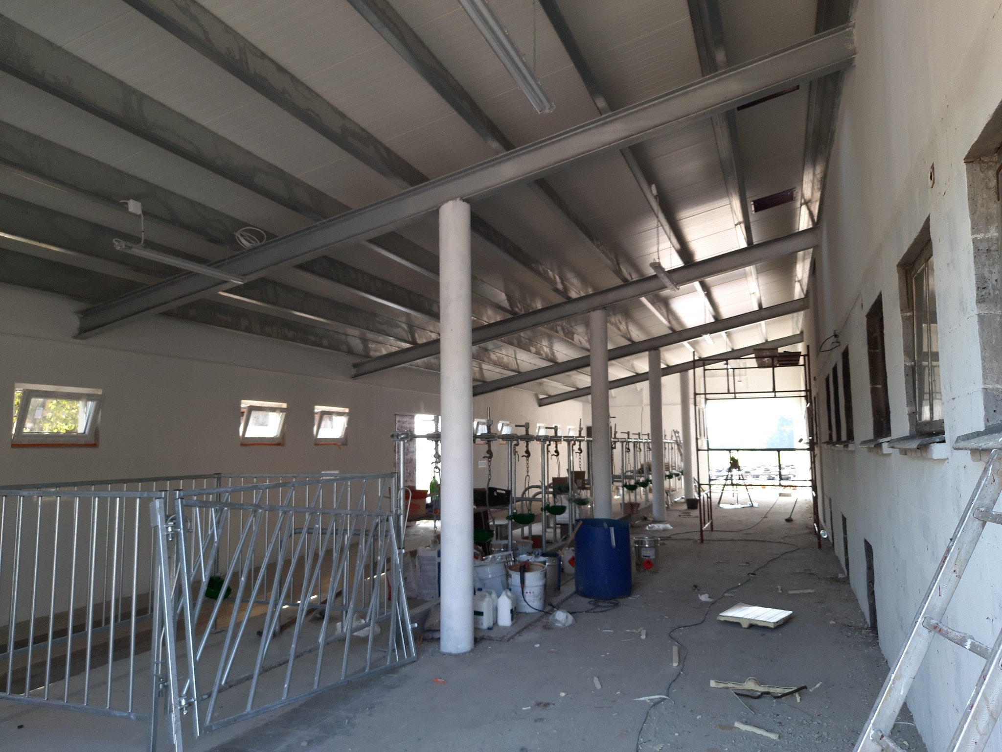 Wnętrze hali w trakcie budowy lub remontu z widoczną konstrukcją dachu z metalowymi belkami, oknami w ścianach i częściowym wyposażeniem.