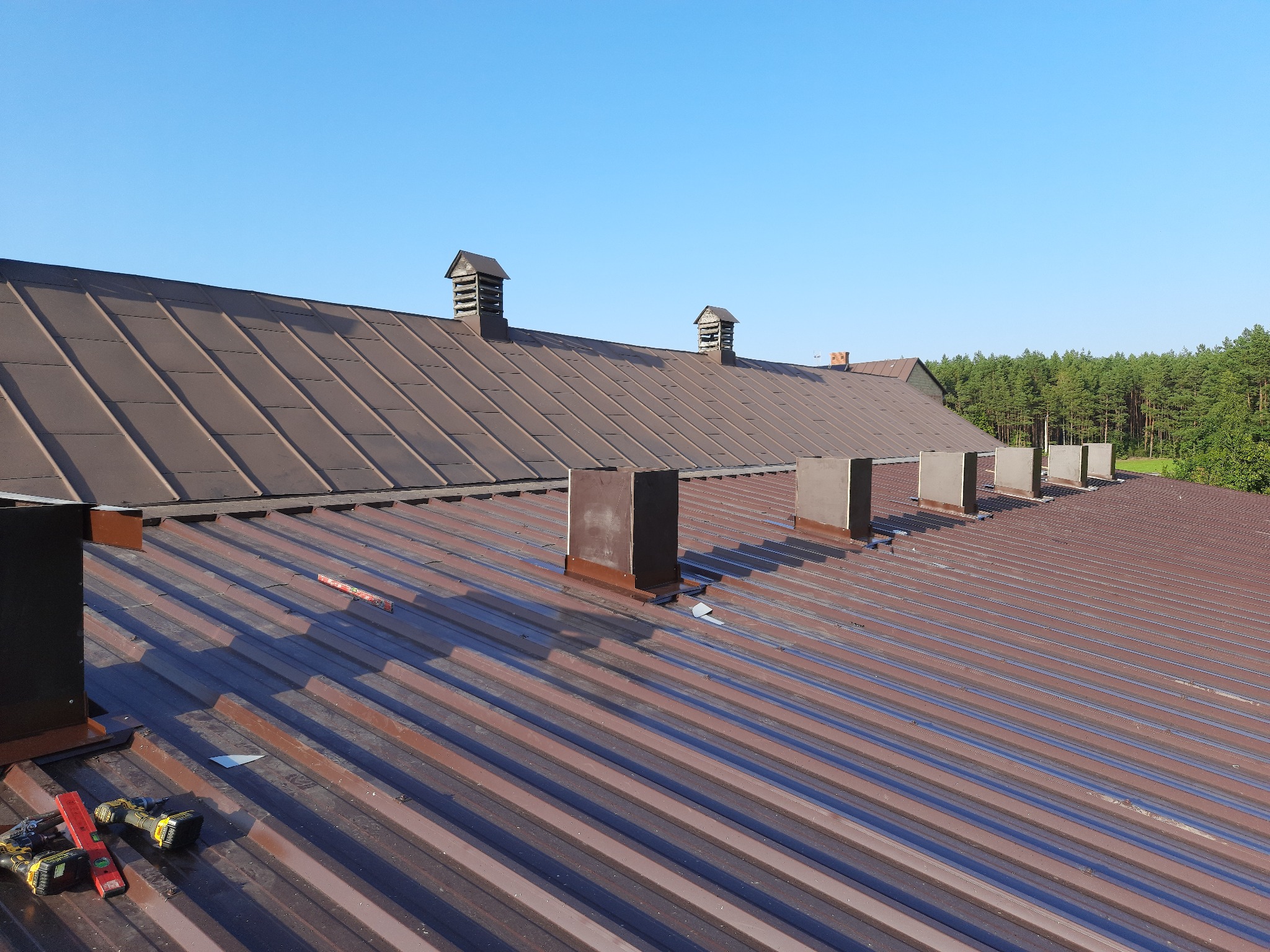 Brązowy dach z blachy trapezowej z kominami wentylacyjnymi, widoczne narzędzia dekarskie i poziomica, w tle las i błękitne niebo.
