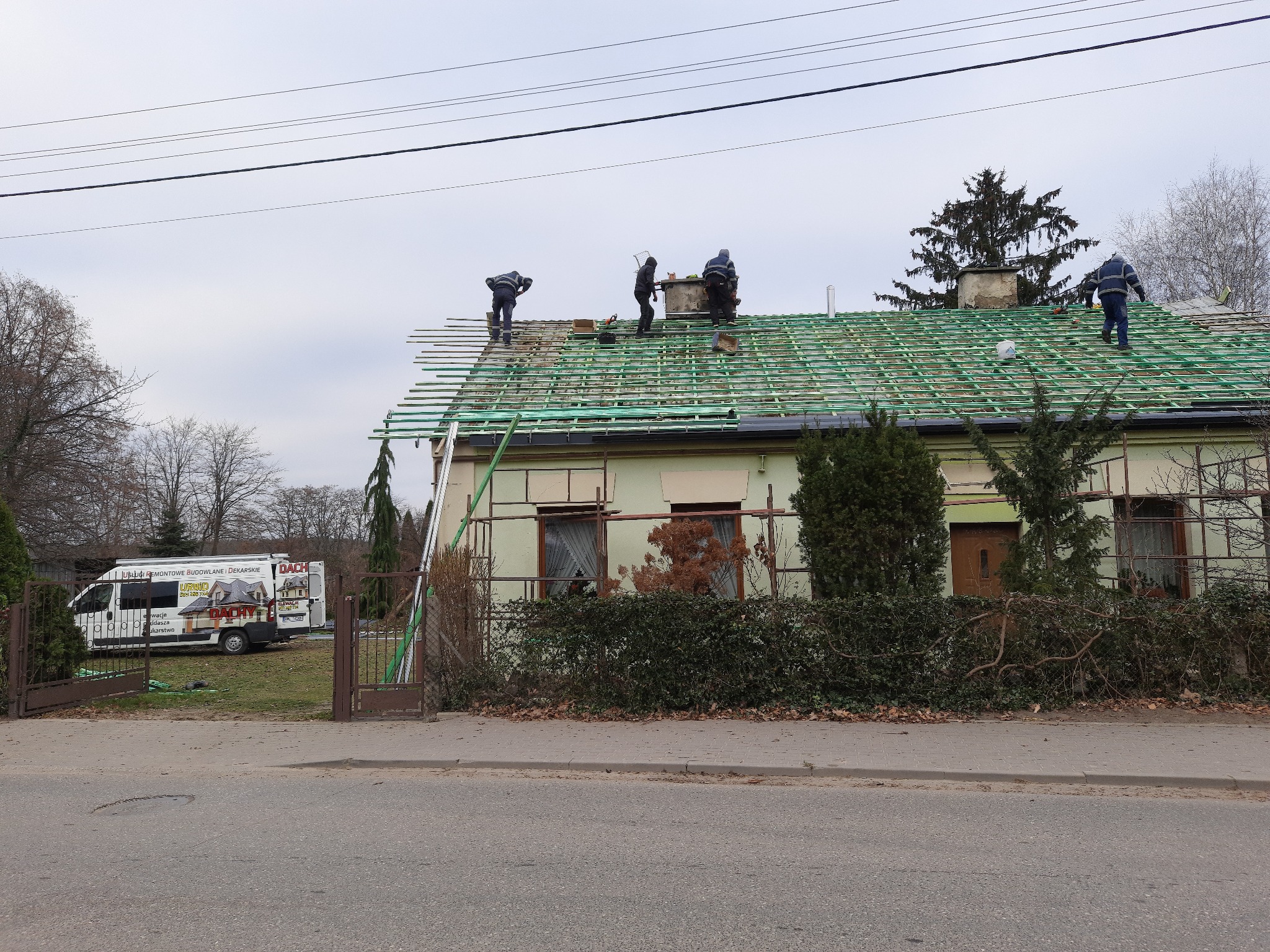 Ekipa dekarzy pracuje na dachu budynku jednorodzinnego, widoczna konstrukcja dachu z łatami, w tle zaparkowany bus firmy dekarskiej.