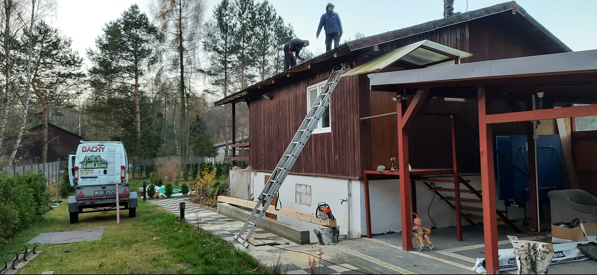 Dwóch dekarzy pracuje na dachu drewnianego domu jednorodzinnego, z widoczną drabiną, piłą łańcuchową, firmowym busem z napisem 'Dachy' i wózkiem z lalką w tle.