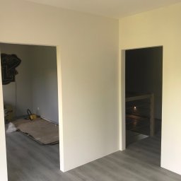 USŁUGI WYKOŃCZENIOWE KRYSTIAN I ŻANETA KASZYŃSCY - Świeżo pomalowane na jasny odcień ściany z dwoma otworami drzwiowymi, widoczne panele podłogowe, w tle w jednym z pomieszczeń widoczny sprzęt i materiały budowlane oraz ozdobna naklejka na ścianie.