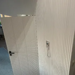 Białe, ryflowane panele ścienne i otwarte drzwi z czarną klamką. Widoczny fragment instalacji domofonowej i gniazdko elektryczne. Nowoczesny design.