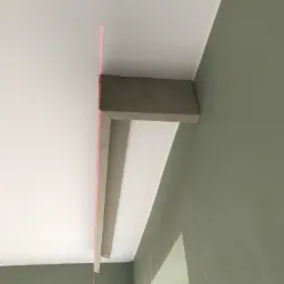 Zamontowana listwa sufitowa z płyty gipsowo-kartonowej, wyznaczona laserem na styku białego sufitu i oliwkowozielonej ściany. Widoczny fragment okna.