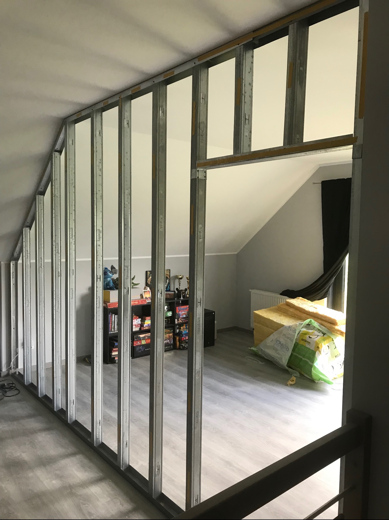 Metalowy szkielet ścianki działowej z profili, widok z góry na poddasze z oknem dachowym, w tle regał z książkami i materiały izolacyjne.