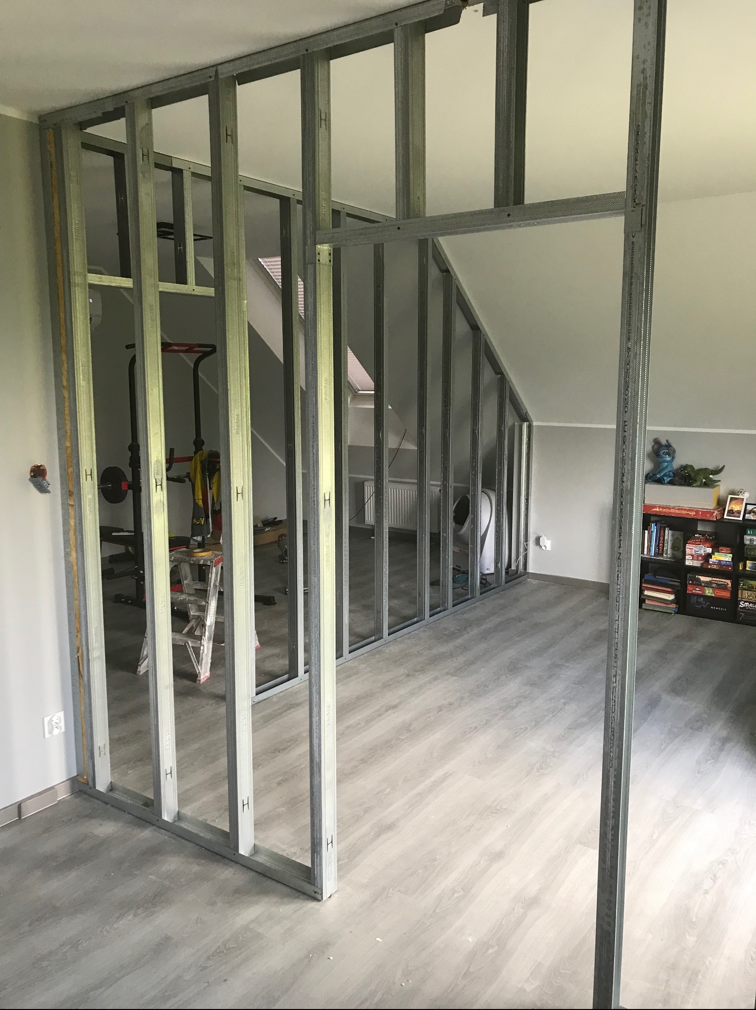 Metalowy szkielet ścianki działowej z profili stalowych w szarym pokoju na poddaszu, widoczne elementy wyposażenia siłowni w tle.