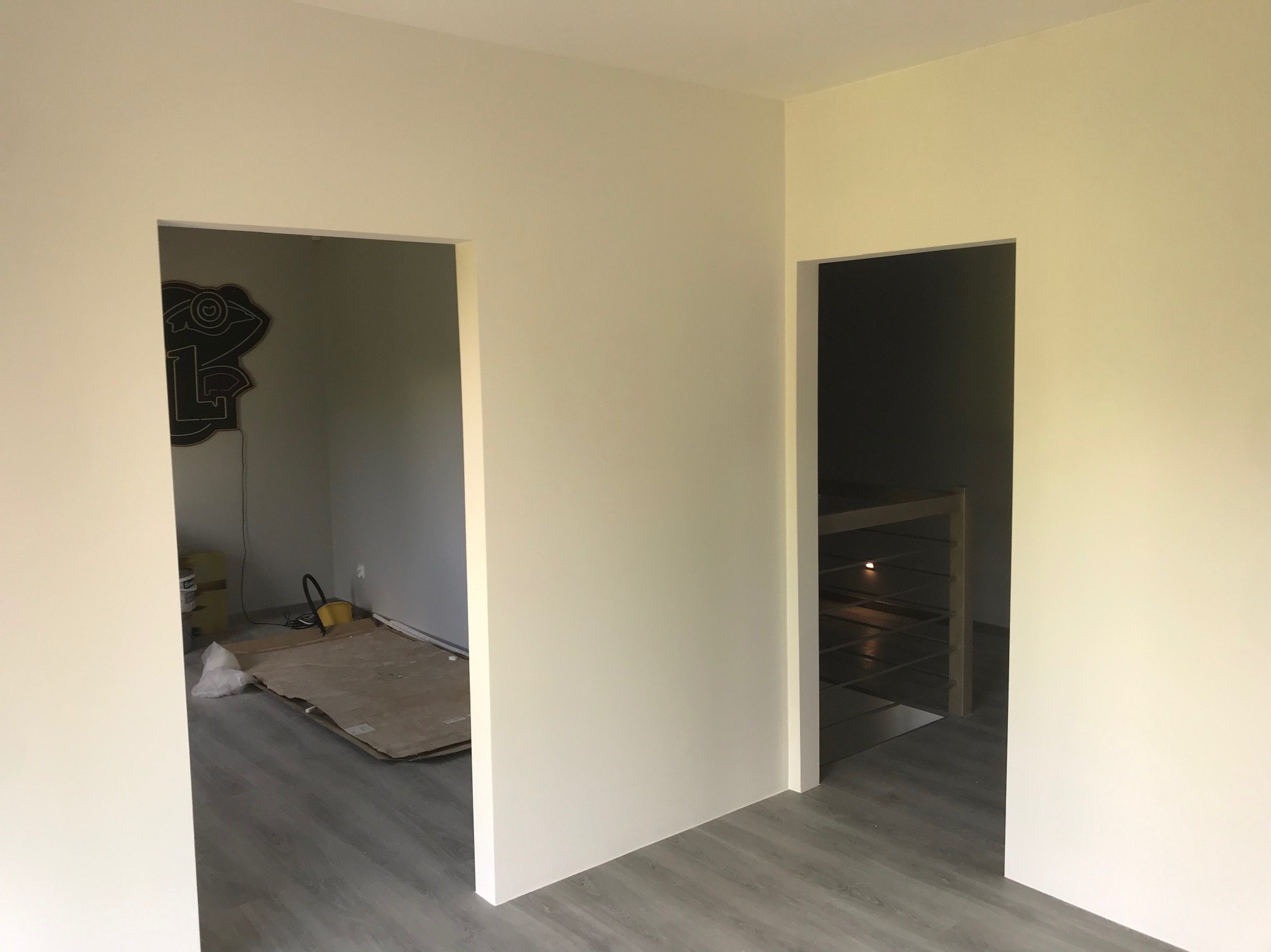 Świeżo pomalowane na jasny odcień ściany z dwoma otworami drzwiowymi, widoczne panele podłogowe, w tle w jednym z pomieszczeń widoczny sprzęt i materiały budowlane oraz ozdobna naklejka na ścianie.