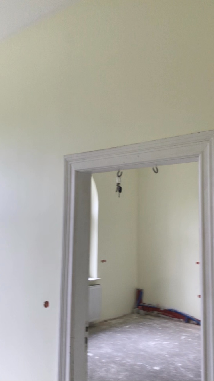 Wnętrze pokoju po malowaniu na jasny kolor, widoczne nowe rury grzewcze i klucze wiszące na framudze drzwi, w tle grzejnik pod oknem.