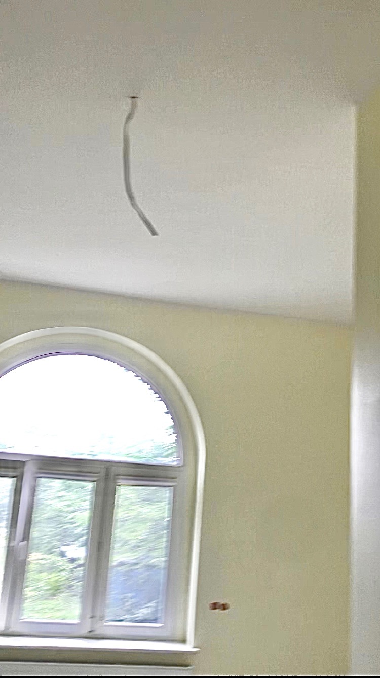 Wnętrze pokoju w trakcie remontu: widoczne fragmenty ściany pomalowanej na jasny kolor, okno z zaokrąglonym łukiem u góry oraz wystający przewód elektryczny z sufitu.