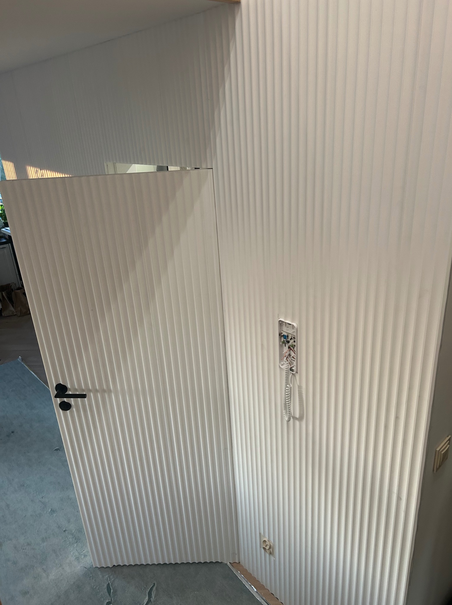 Białe, ryflowane panele ścienne i otwarte drzwi z czarną klamką. Widoczny fragment instalacji domofonowej i gniazdko elektryczne. Nowoczesny design.