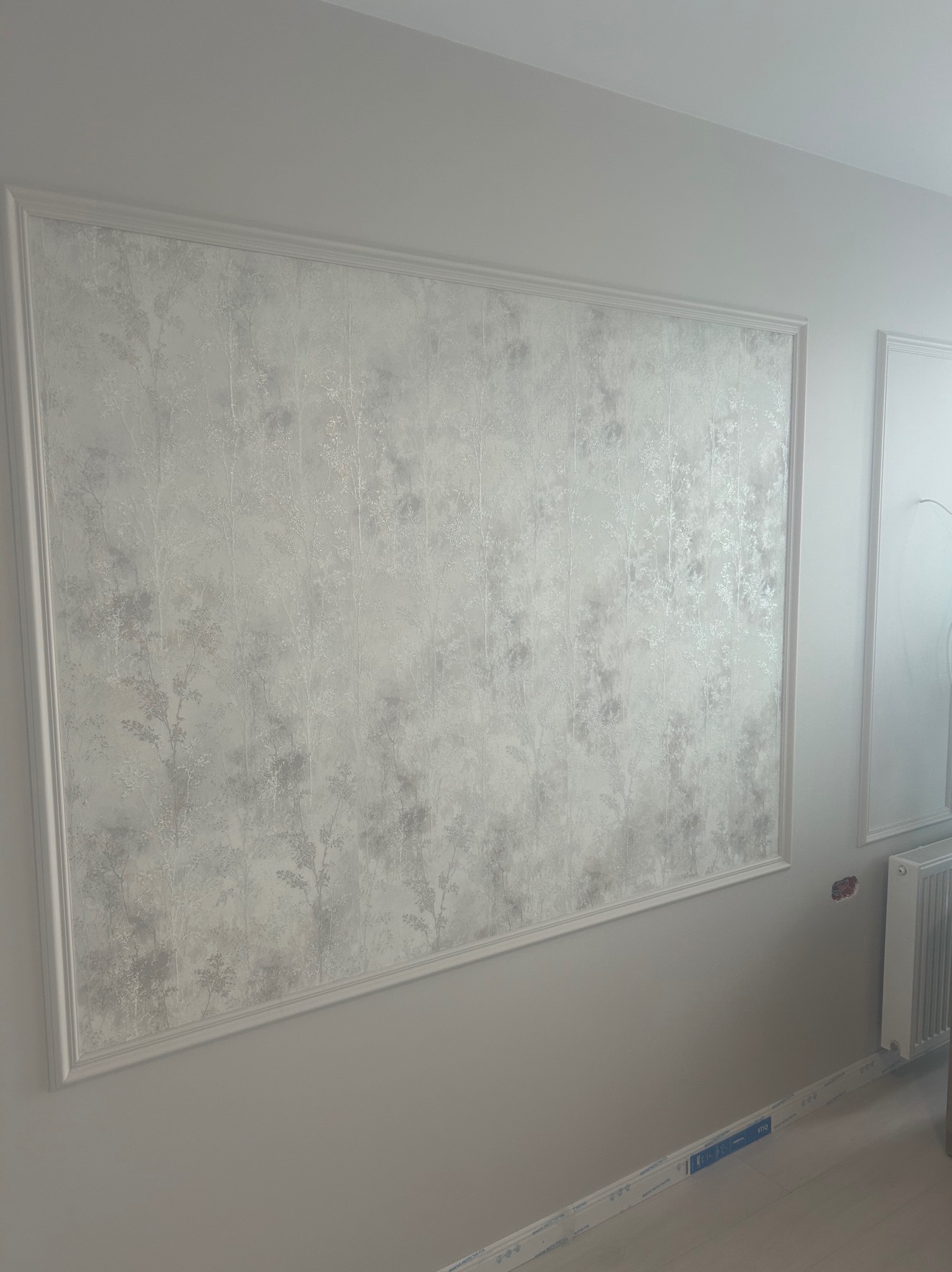 Elegancka ściana z dekoracyjną ramą i tapetą w subtelny wzór drzew, uzupełniona o listwy przypodłogowe i fragment grzejnika. Jasne, stonowane barwy.