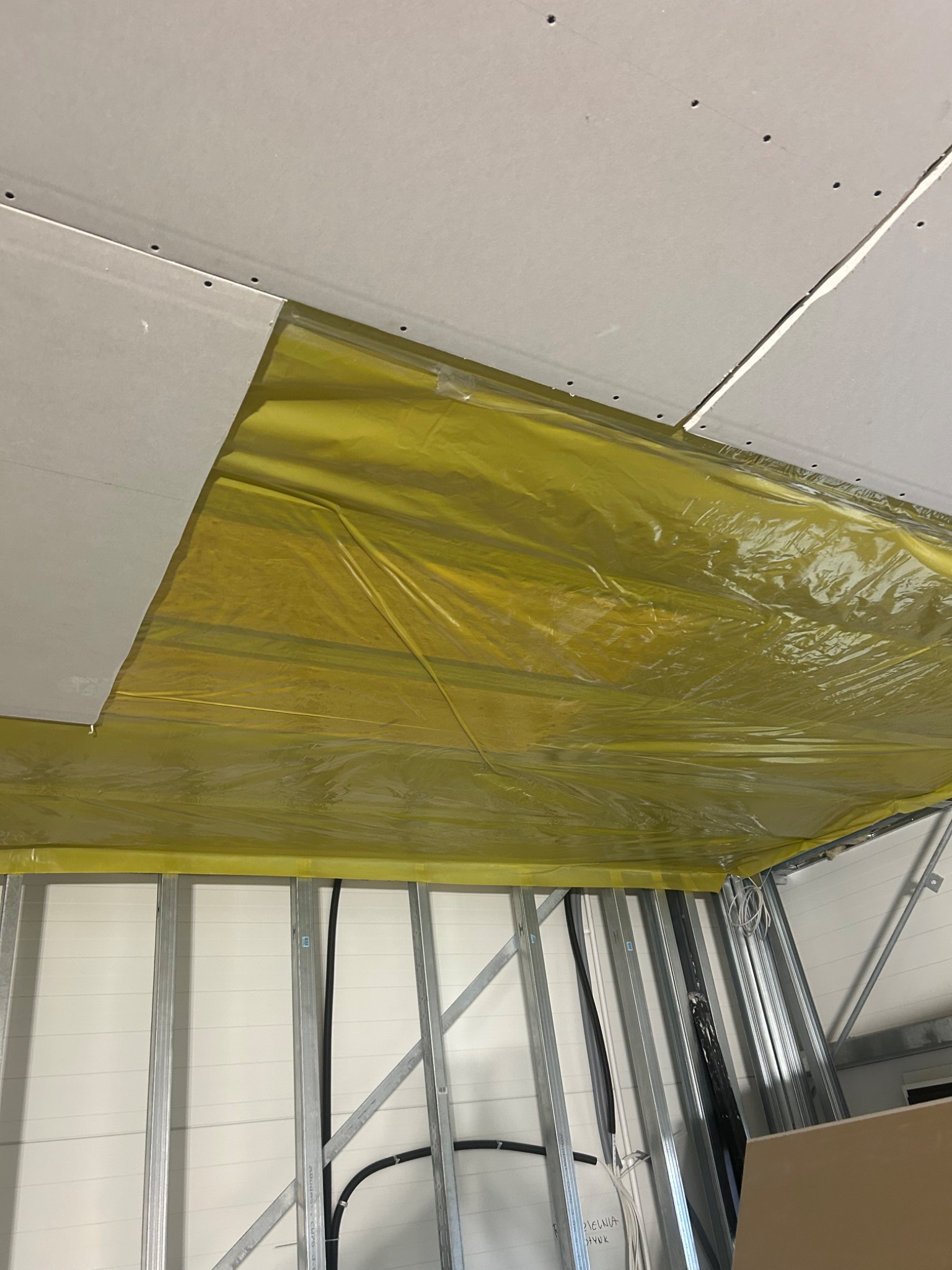 Montaż płyty G-K na metalowym stelażu z folią paroizolacyjną. Widoczne kable i przewody. Ujęcie z dołu, sufit w trakcie prac.