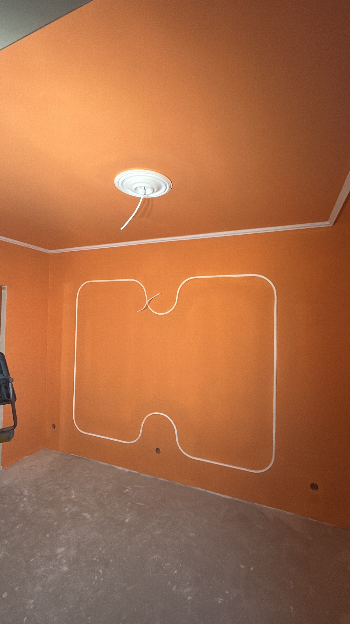 Pomarańczowe wnętrze w trakcie wykańczania: ściany z dekoracyjną listwą, sufit z rozetą i wystającymi kablami, surowa podłoga. Widoczne gniazdka elektryczne.