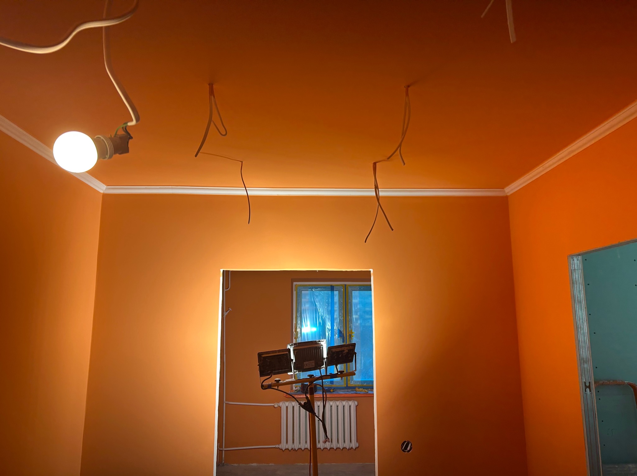 Pomarańczowe wnętrze po malowaniu z widocznymi kablami i lampą halogenową. Świeżo pomalowane ściany z białym wykończeniem sufitu. Widok na okno zza ściany.