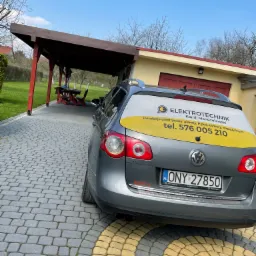 Szary samochód marki Volkswagen z naklejoną reklamą firmy Elektrotechnik Kamil Mondzelewski na tylnej szybie, zaparkowany na brukowanym podjeździe przed garażem i zadaszeniem.