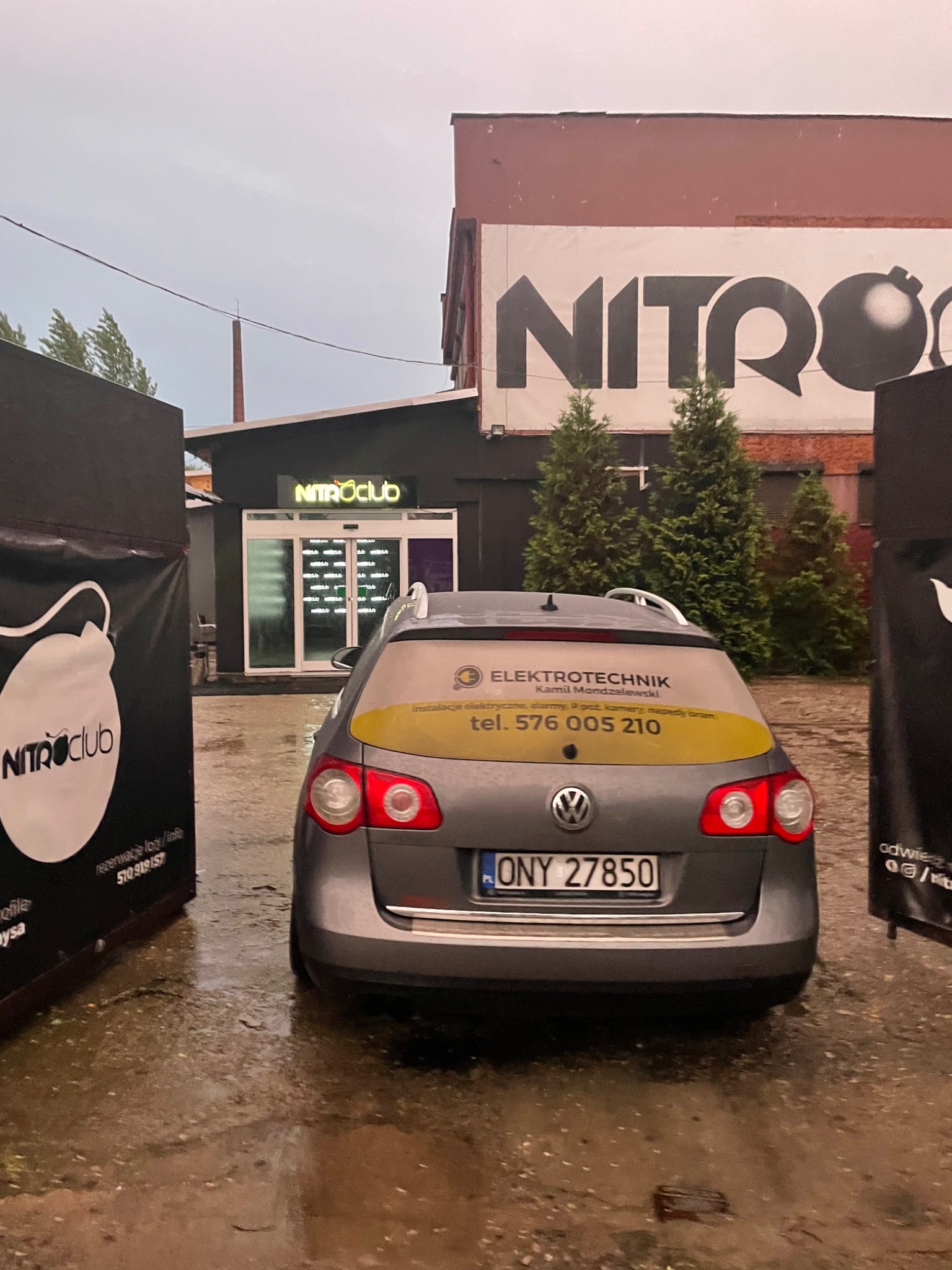 Szary samochód osobowy z naklejką reklamową firmy Elektrotechnik Kamil Mondzelewski na tylnej szybie, zaparkowany na mokrym placu przed budynkiem NitroClub z neonowym szyldem.