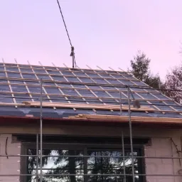 Dach w trakcie wymiany: widoczna membrana dachowa, łaty, rusztowanie i fragment elewacji budynku w tle, wieczorne światło.