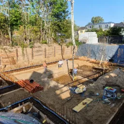 Wykop pod fundamenty domu w Opolu, z widocznym zbrojeniem, szalunkiem, rurami kanalizacyjnymi i pracownikami. Na pierwszym planie kubek po kawie z logo firmy.