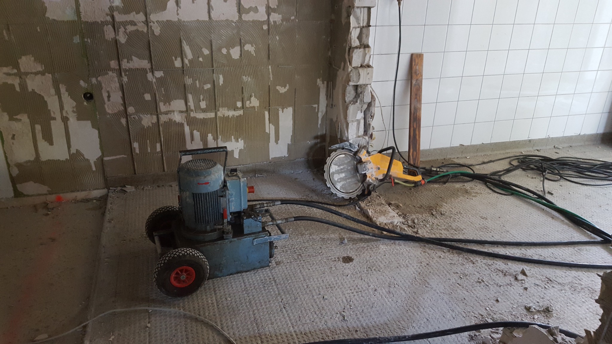 Hydrauliczna piła do betonu z tarczą diamentową przecina ścianę w trakcie remontu, widoczny agregat hydrauliczny i plątanina kabli na podłodze.