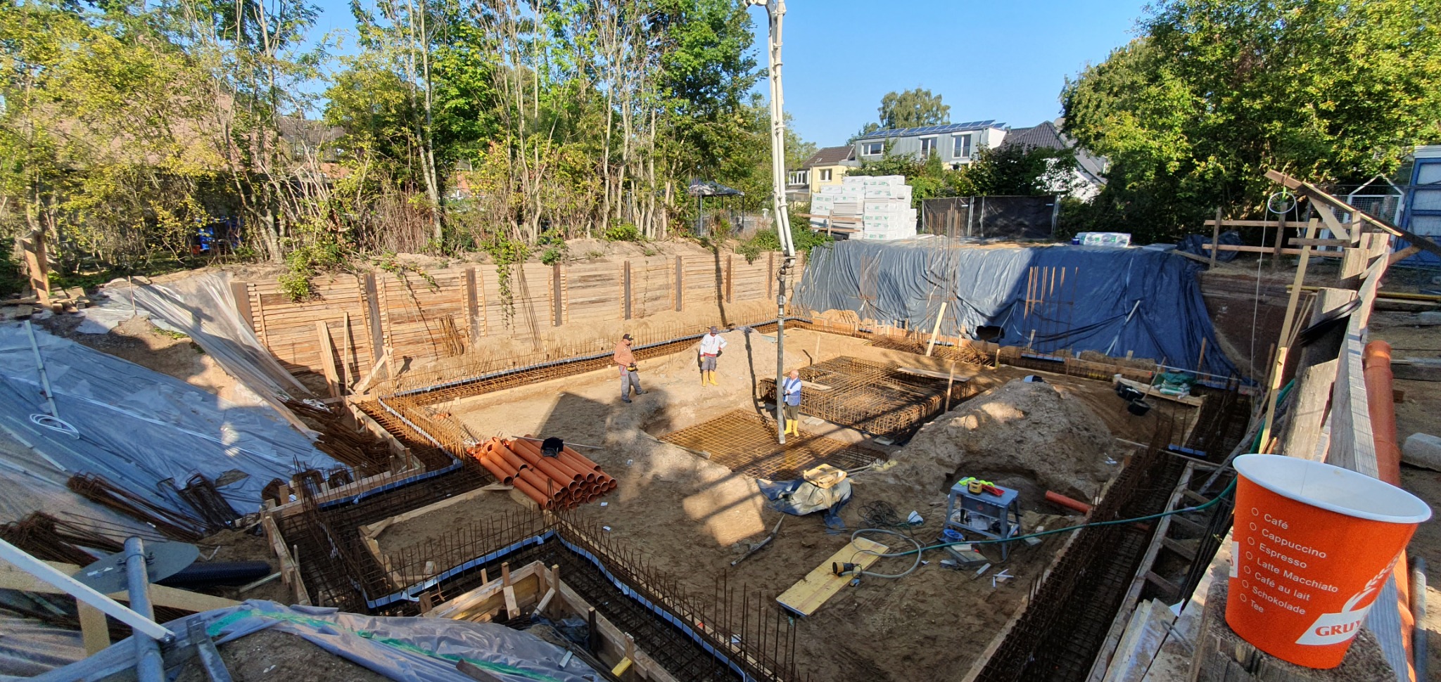 Wykop pod fundamenty domu w Opolu, z widocznym zbrojeniem, szalunkiem, rurami kanalizacyjnymi i pracownikami. Na pierwszym planie kubek po kawie z logo firmy.