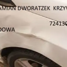 Biały samochód z wgnieceniem na karoserii i napisem 'AUTONAPRAWA DAMIAN DWORZETZEK KRZYWE KOLANO 14' oraz numerem telefonu, wskazujący na ofertę usług naprawy pojazdów.