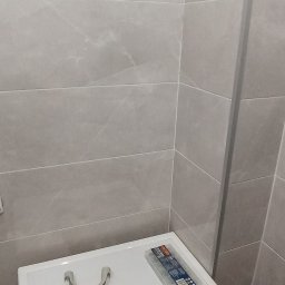 Daniel Kasprzak - Fragment łazienki z szarymi płytkami, na umywalce leży uchwyt rury i pudełko z kołkami montażowymi, zasłonięte palcem.