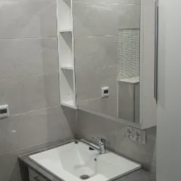 Nowoczesna łazienka w szarościach: biała umywalka nablatowa z szafką i pralką, lustro z półkami, płytki imitujące marmur, dekoracyjna mozaika w odbiciu.