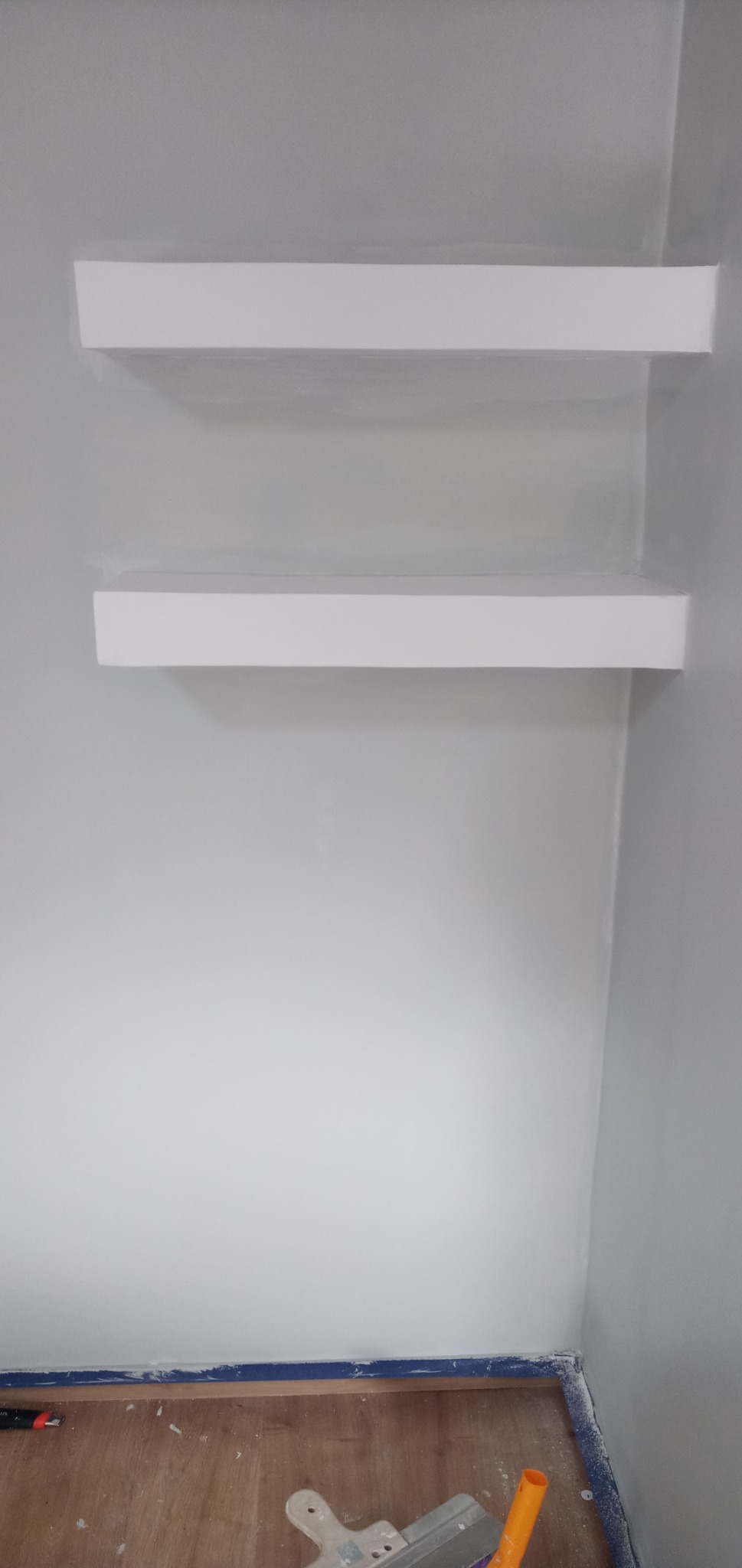 Dwie białe półki zamontowane na świeżo pomalowanej szarej ścianie, widoczne ślady taśmy malarskiej na krawędziach i narzędzia malarskie na podłodze z paneli.