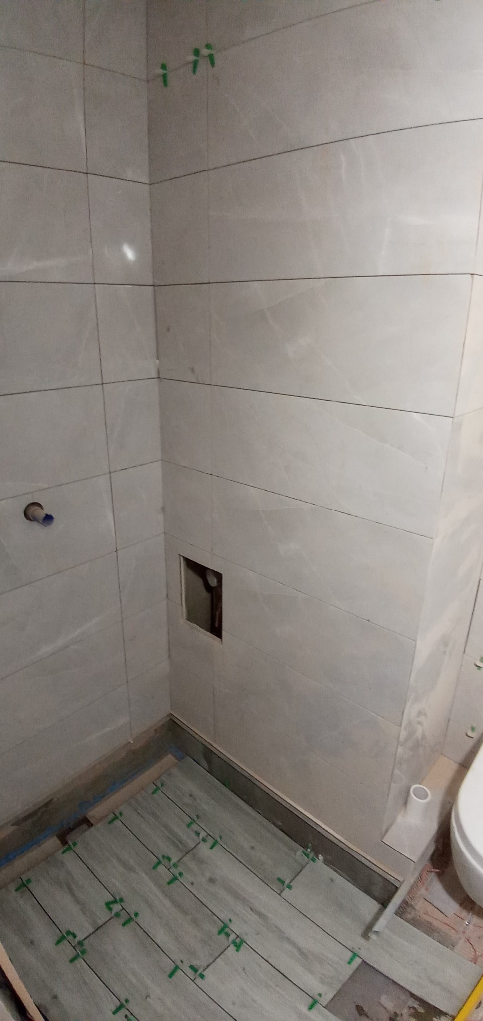 Łazienka w trakcie wykańczania: ułożone płytki podłogowe imitujące drewno z zielonymi krzyżykami dystansowymi, ściany wyłożone płytkami imitującymi marmur, widoczny otwór instalacyjny.