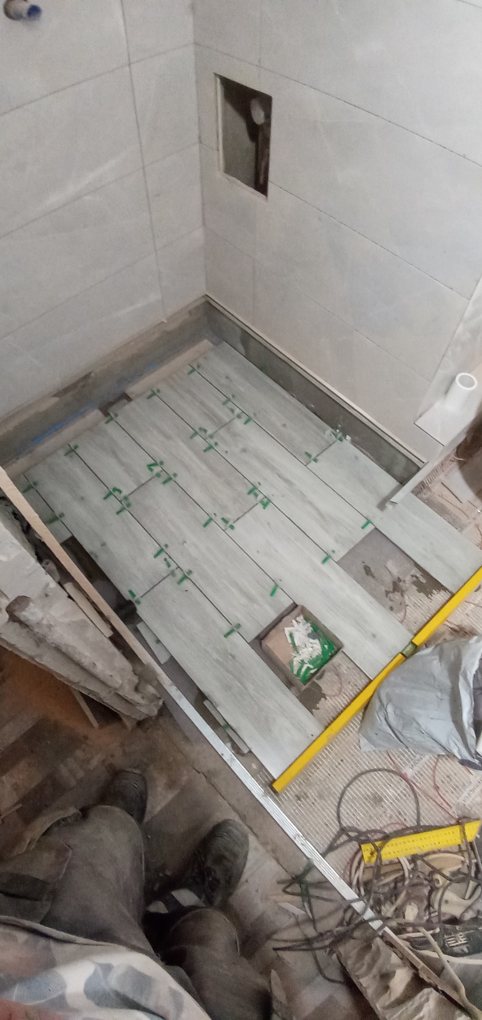 Fragment łazienki w trakcie remontu: ułożone częściowo płytki podłogowe imitujące drewno, zielone krzyżyki dystansowe, poziomica, widoczne narzędzia i materiały budowlane.
