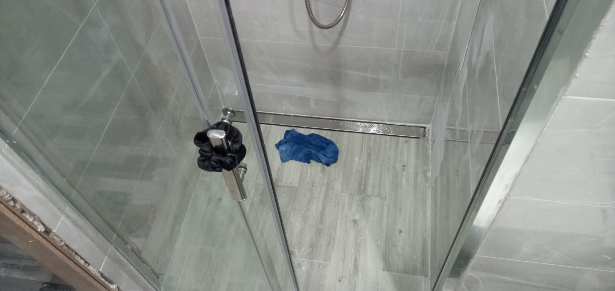 Szklana kabina prysznicowa z szarymi płytkami, czarną gumką do włosów na uchwycie i niebieską ścierką na podłodze, widok z góry.