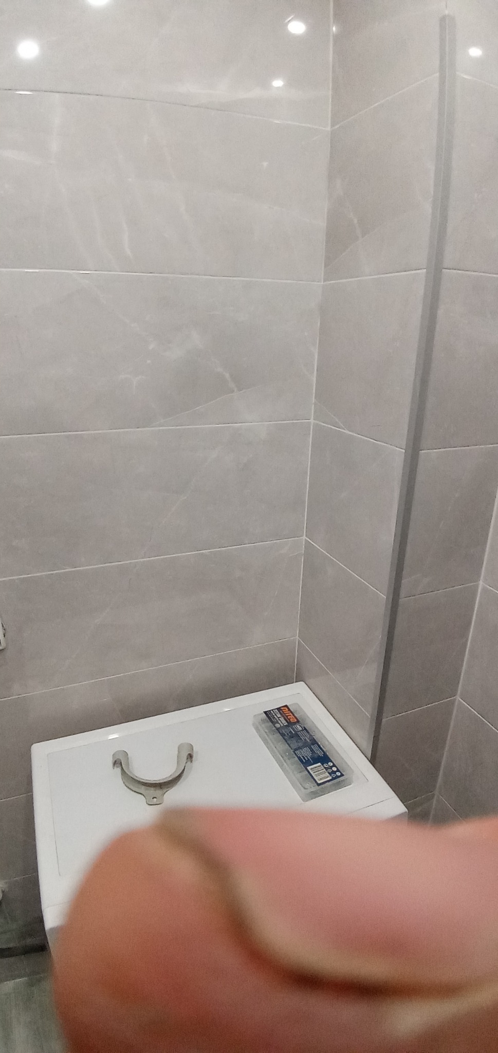 Fragment łazienki z szarymi płytkami, na umywalce leży uchwyt rury i pudełko z kołkami montażowymi, zasłonięte palcem.