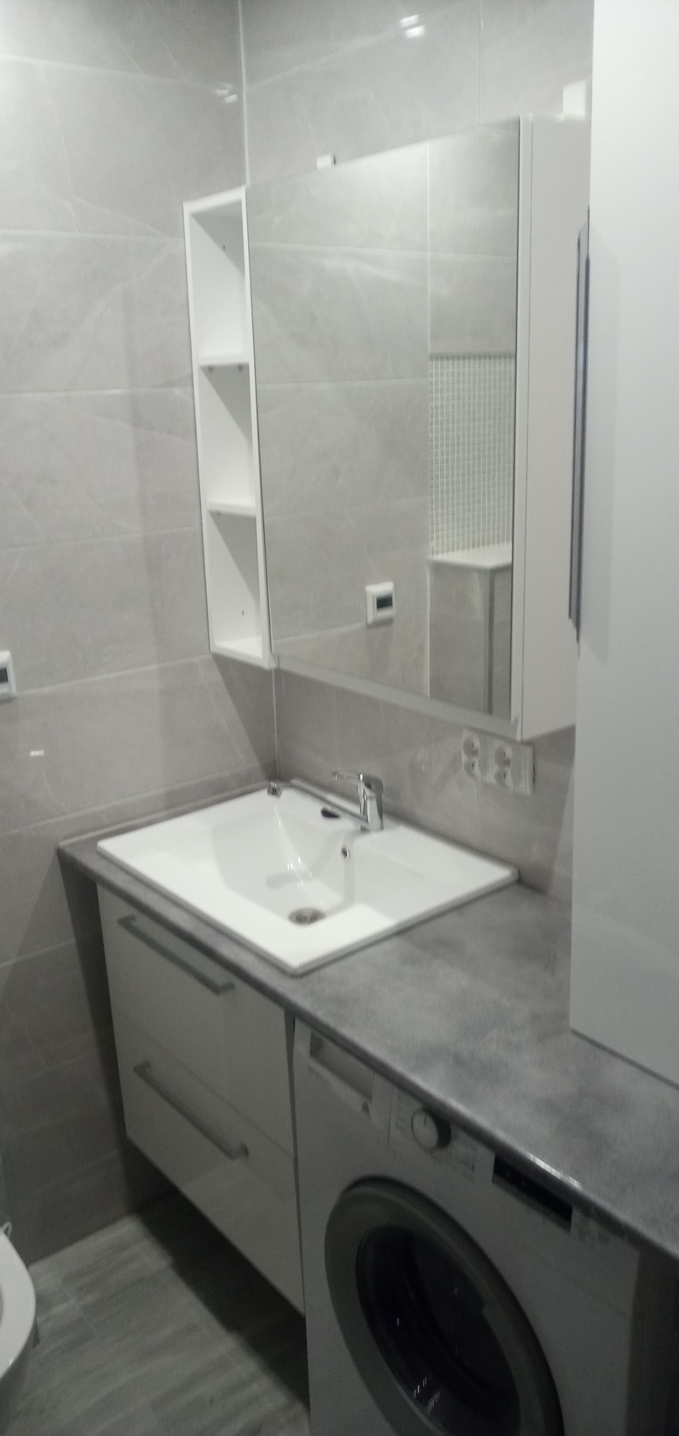 Nowoczesna łazienka w szarościach: biała umywalka nablatowa z baterią, szafka pod umywalką z szufladami, lustro z szafką, białe półki, pralka pod blatem, szare płytki na ścianach i podłodze.