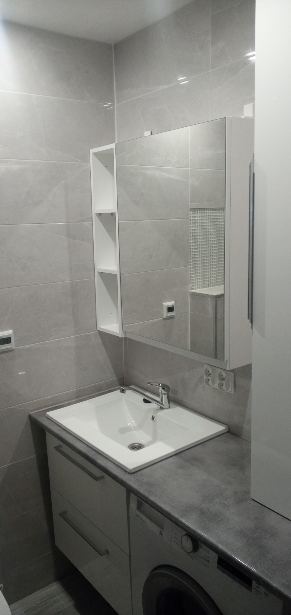 Nowoczesna łazienka w szarościach: biała umywalka nablatowa z szafką i pralką, lustro z półkami, płytki imitujące marmur, dekoracyjna mozaika w odbiciu.