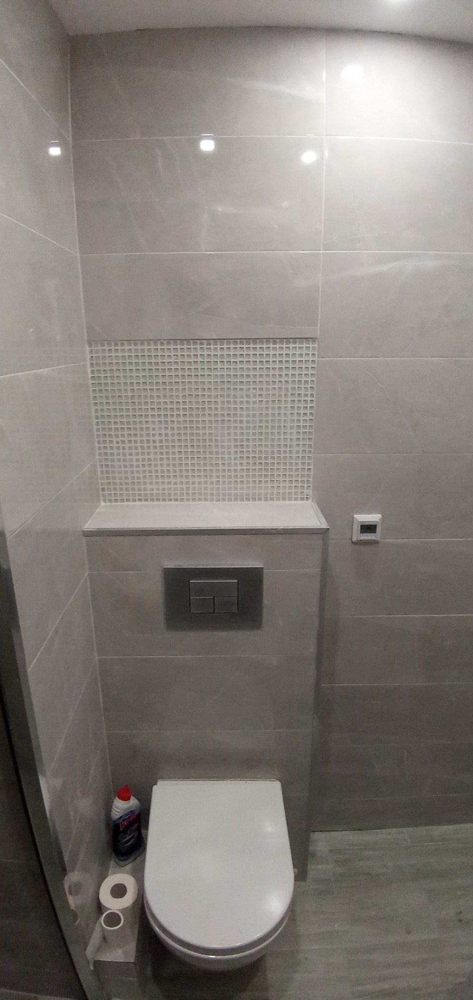 Szara łazienka z mozaiką nad zabudową toalety, widoczny przycisk spłuczki, termostat i akcesoria toaletowe.