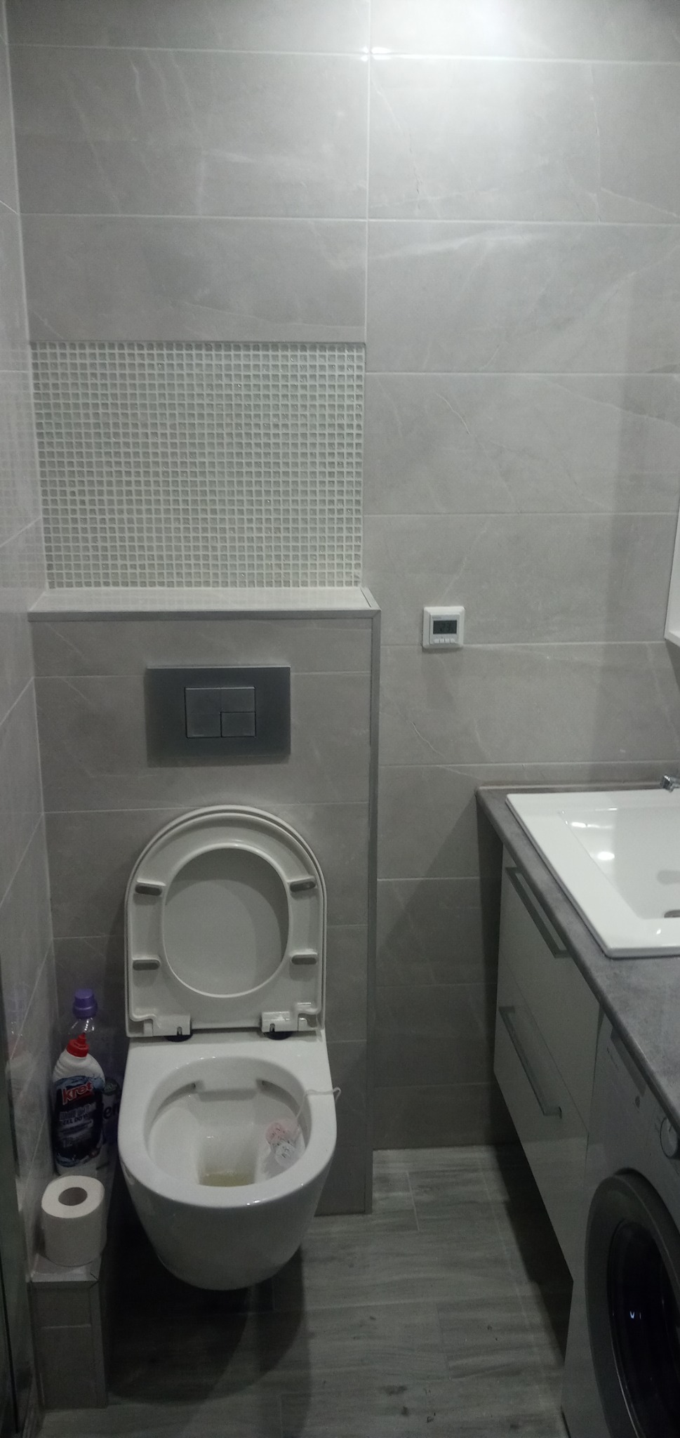 Szara łazienka w Toruniu po remoncie: wisząca toaleta z otwartą klapą, mozaika nad zabudową stelaża, umywalka z szafką i pralka.