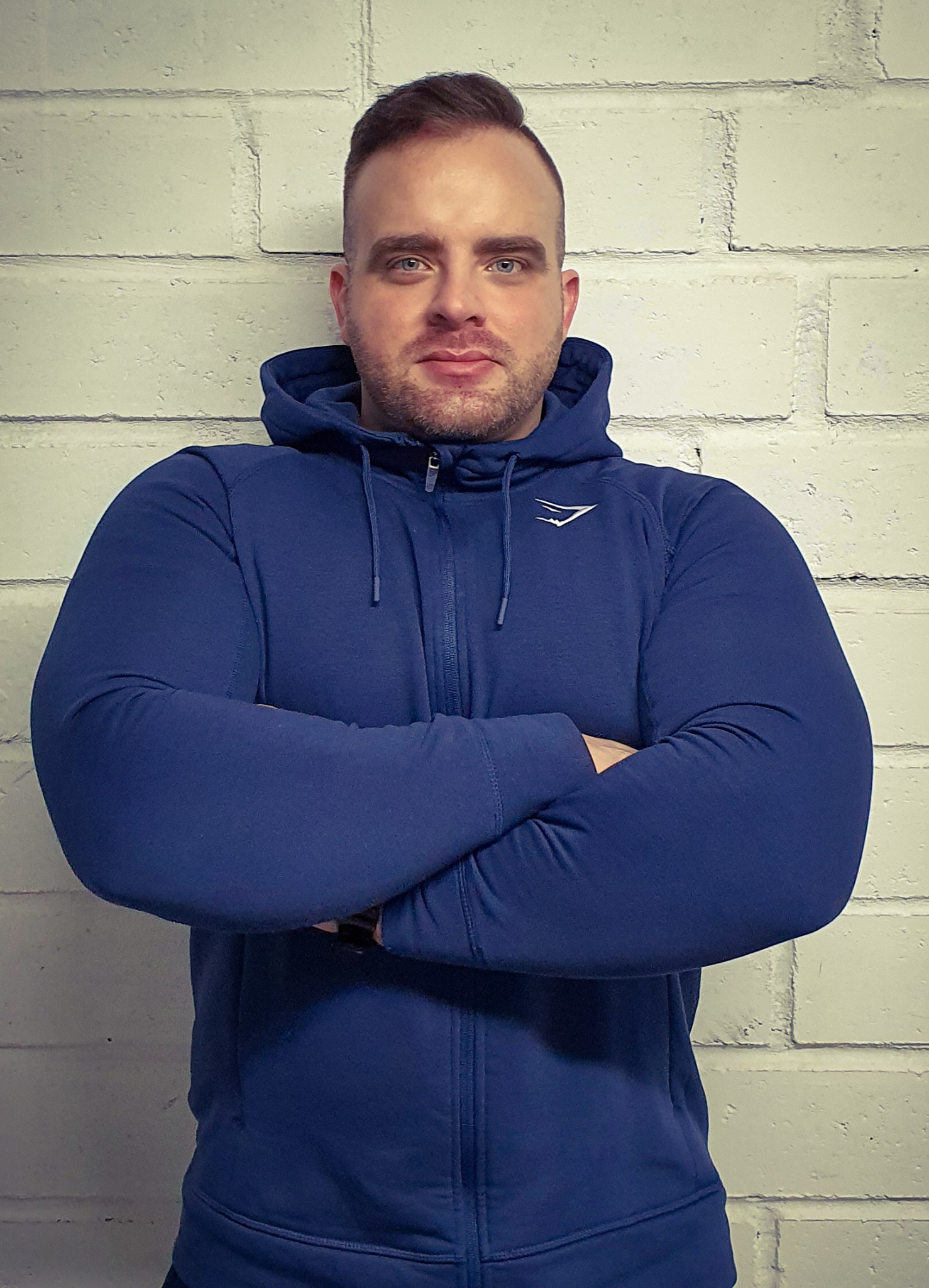 Portret mężczyzny w niebieskiej bluzie z kapturem, z założonymi rękami na tle białej ceglanej ściany.