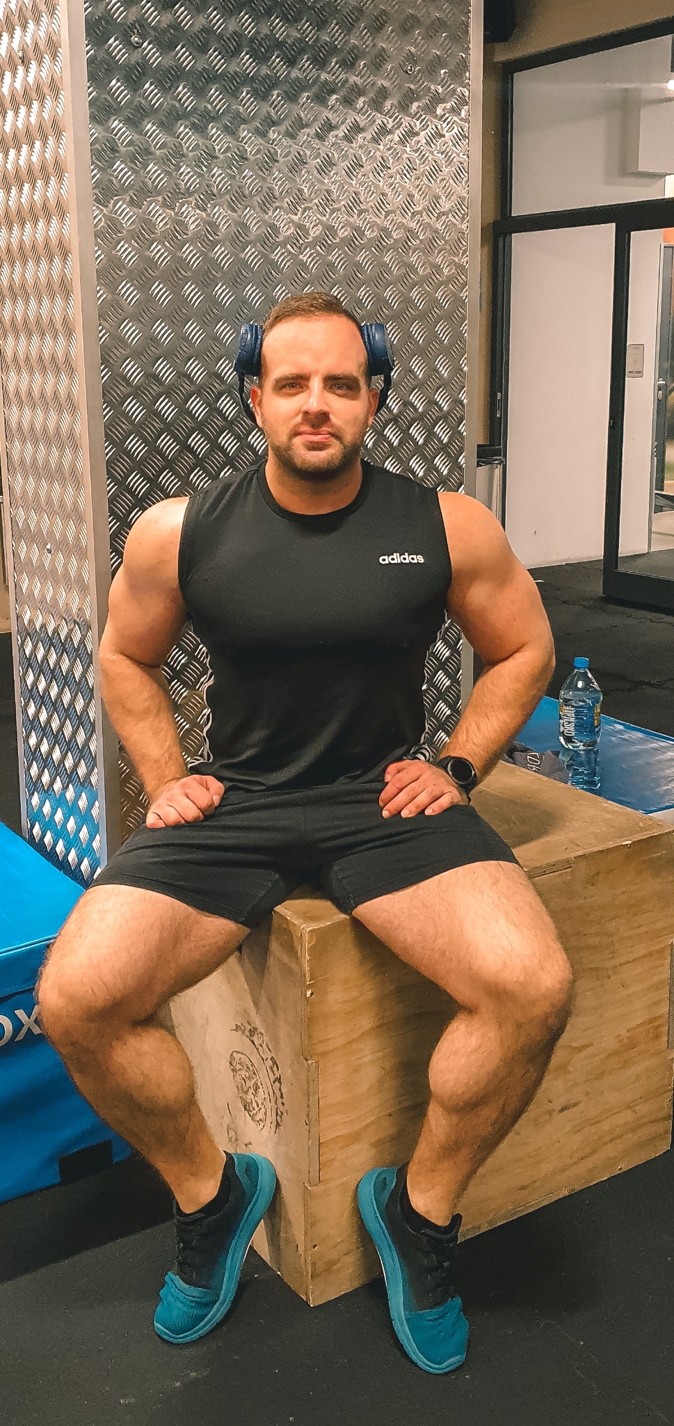 Mężczyzna w sportowym stroju i słuchawkach siedzi na drewnianej skrzyni w siłowni, w tle butelka wody.