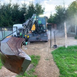 Henbud - Żółto-czarna koparka Volvo z łyżką w pierwszym planie, myta pod ciśnieniem na żwirowej powierzchni, widoczna para wodna, w tle ogrodzenie z siatki i drzewa.