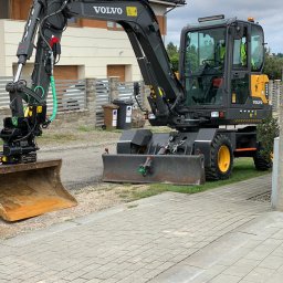 Henbud - Żółto-czarna koparka kołowa Volvo EW60E z dwoma łyżkami, zaparkowana na kostce brukowej przed domem z szarą dachówką.