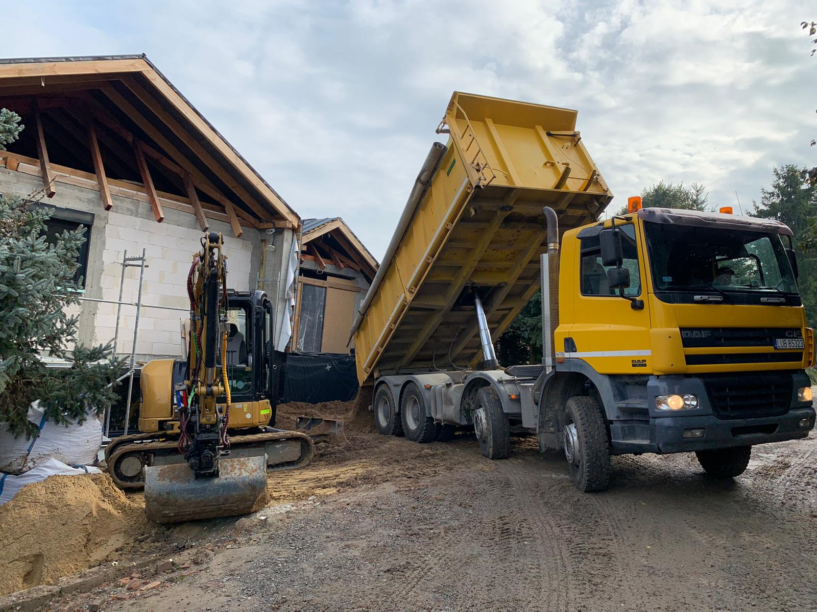 Żółta wywrotka DAF wysypuje ziemię obok budynku w stanie surowym otwartym, obok stoi mała koparka, widoczne ślady opon na błotnistym terenie.
