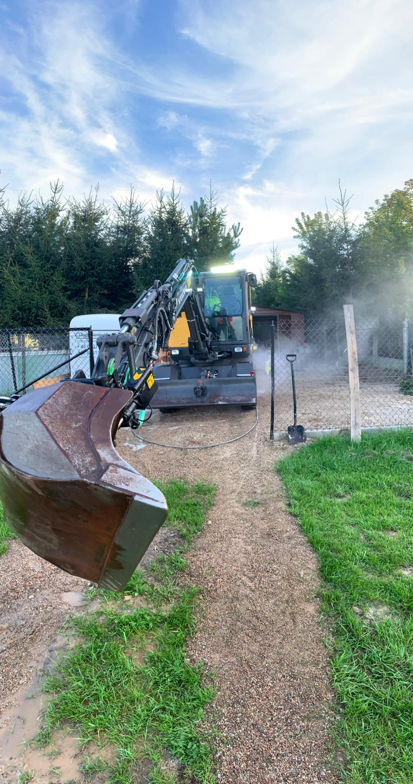 Żółto-czarna koparka Volvo z łyżką w pierwszym planie, myta pod ciśnieniem na żwirowej powierzchni, widoczna para wodna, w tle ogrodzenie z siatki i drzewa.