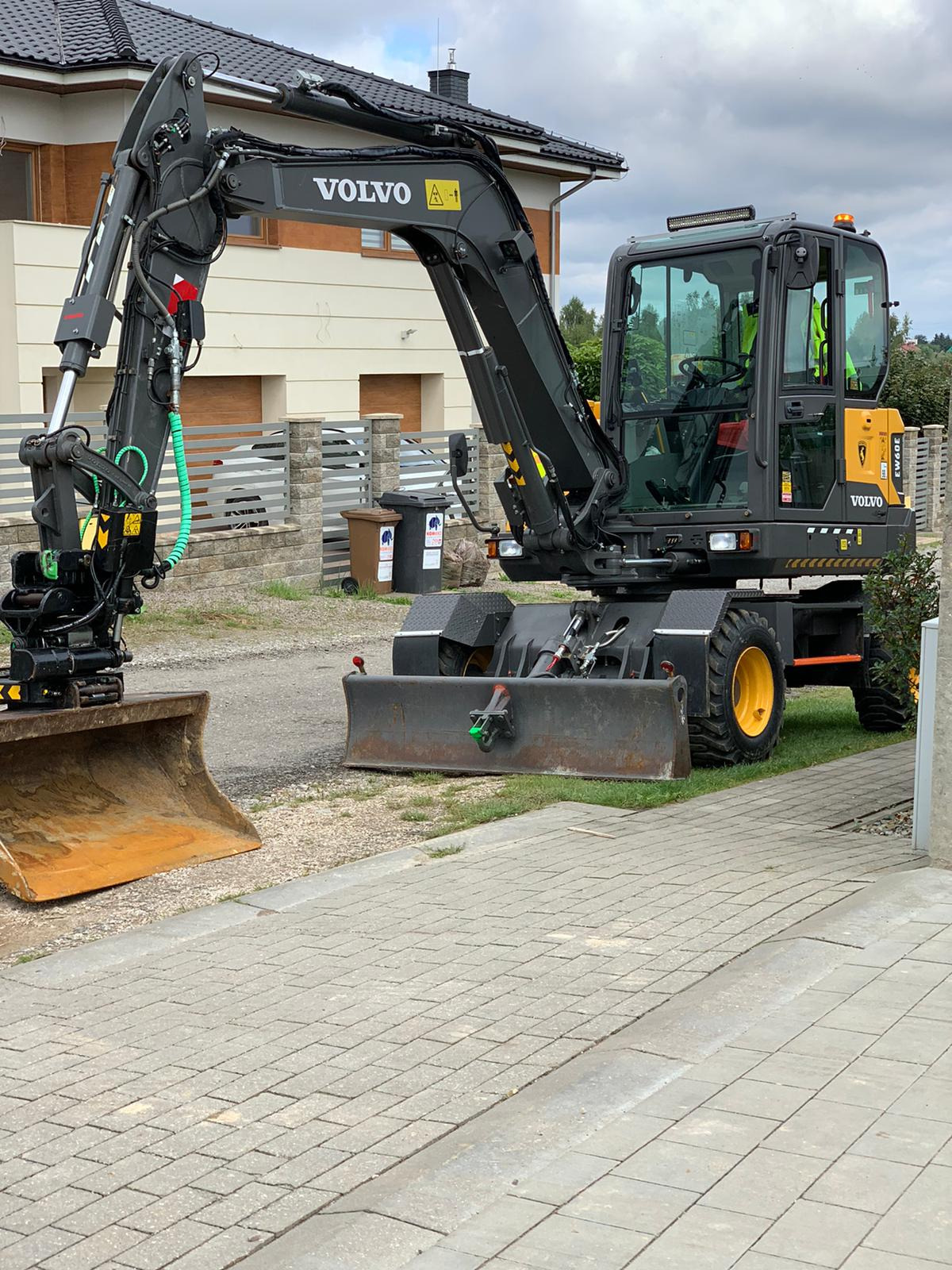 Żółto-czarna koparka kołowa Volvo EW60E z dwoma łyżkami, zaparkowana na kostce brukowej przed domem z szarą dachówką.