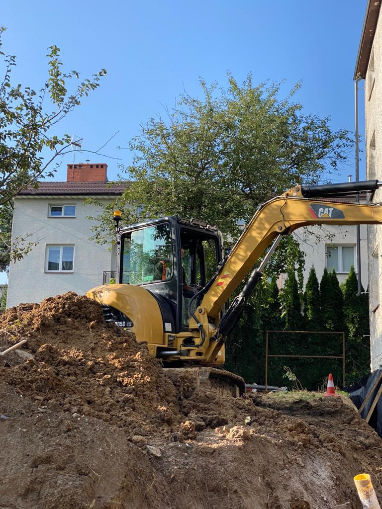 Żółta minikoparka CAT 305C częściowo zagłębiona w wykopie ziemnym obok budynku mieszkalnego, widoczne drzewa i niebieskie niebo.