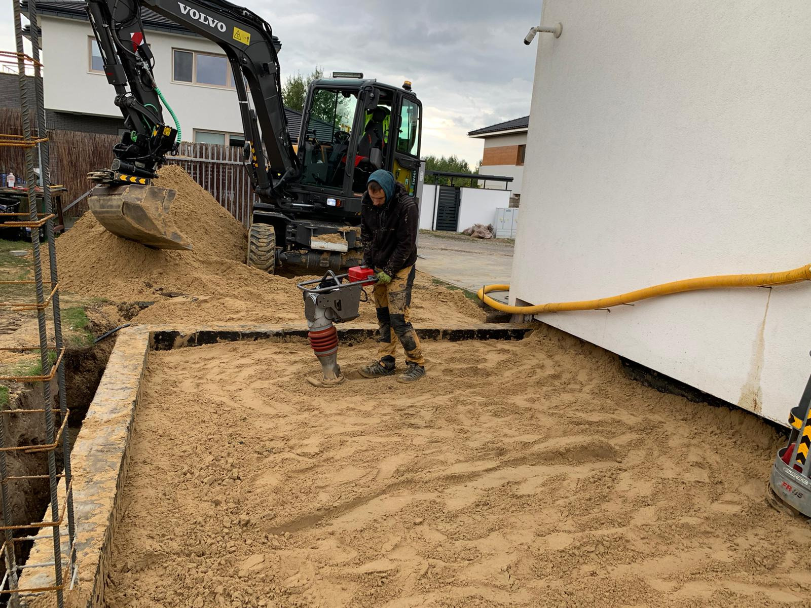 Operator zagęszcza piasek ubijarką skoczną obok fundamentów domu w Lublinie, na drugim planie czarna koparka Volvo z łyżką pełną piachu.