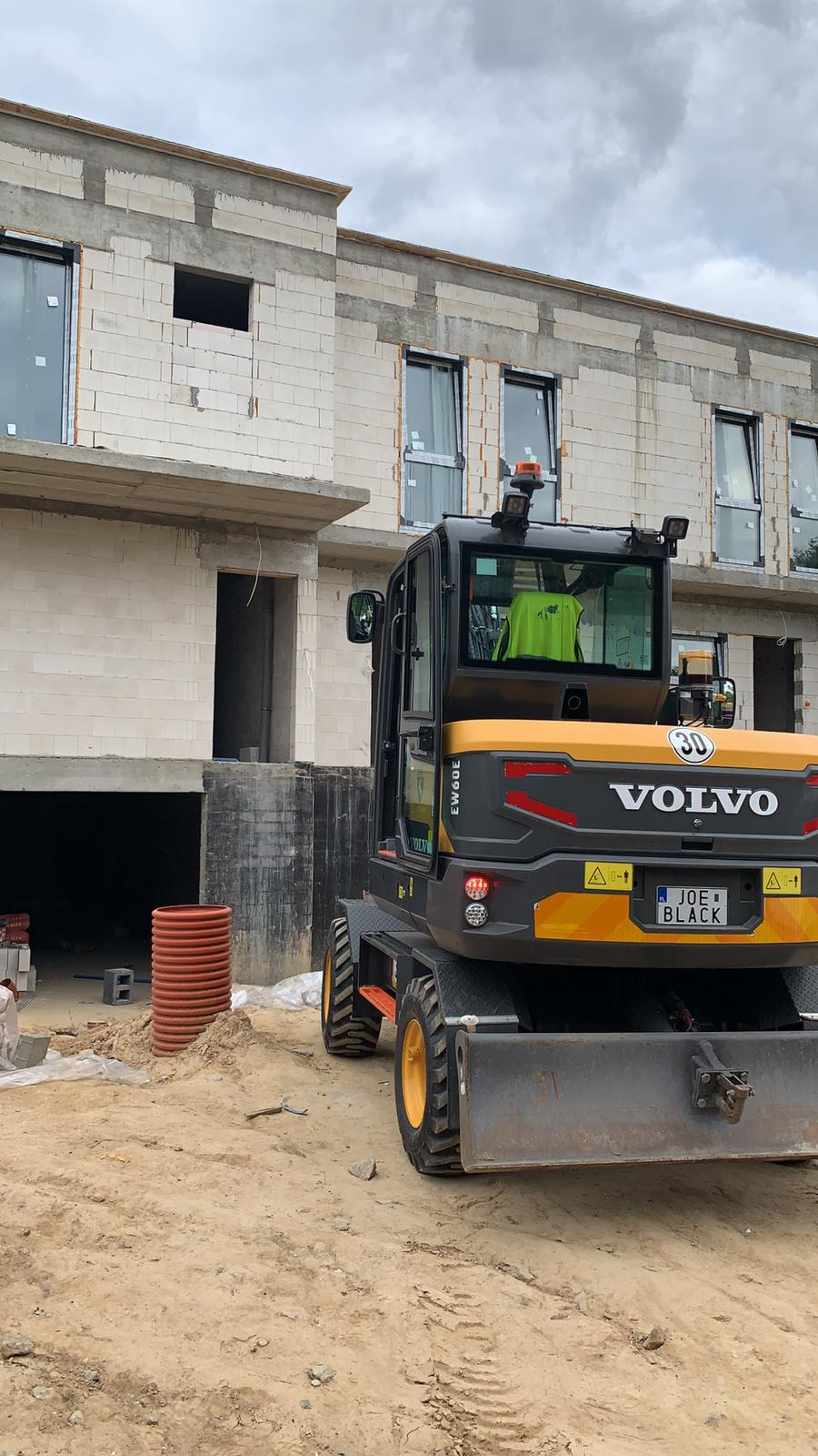 Żółto-czarna koparka Volvo EW60E z widoczną tablicą rejestracyjną 'JOE BLACK' na tle surowego budynku w trakcie budowy z białego bloczka.