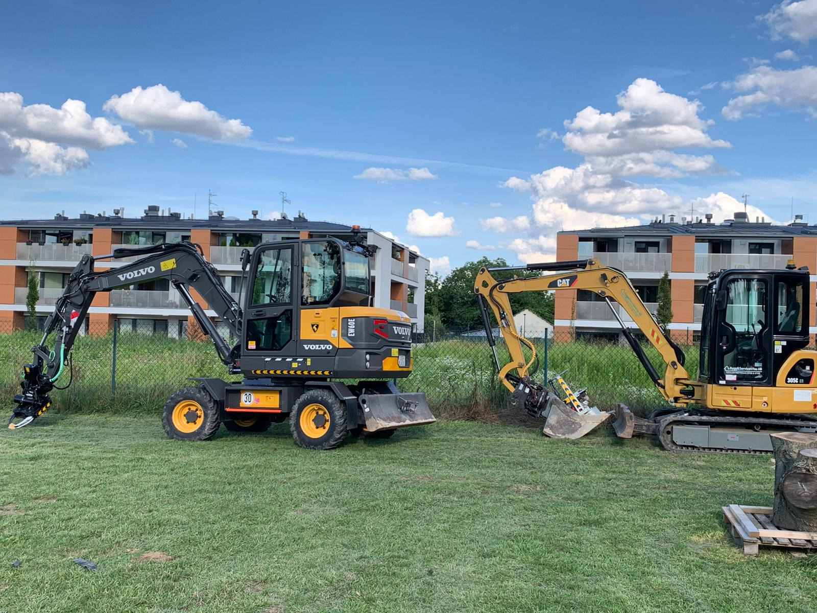 Dwie minikoparki, Volvo i Caterpillar, stoją na trawniku przed budynkiem mieszkalnym w słoneczny dzień. Koparka Volvo ma zamontowany frez do pni drzew, a Caterpillar łyżkę. Obie maszyny gotowe...