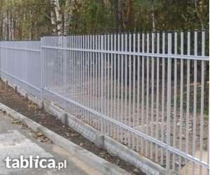 Szary metalowy płot sztachetowy z poziomą poprzeczką, oddzielający chodnik od trawnika, widoczny z boku.
