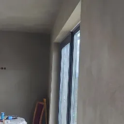 Wnętrze z tynkami maszynowymi w odcieniach szarości, widoczne niedokończone podłączenie oświetlenia i okno z widokiem na zewnątrz.