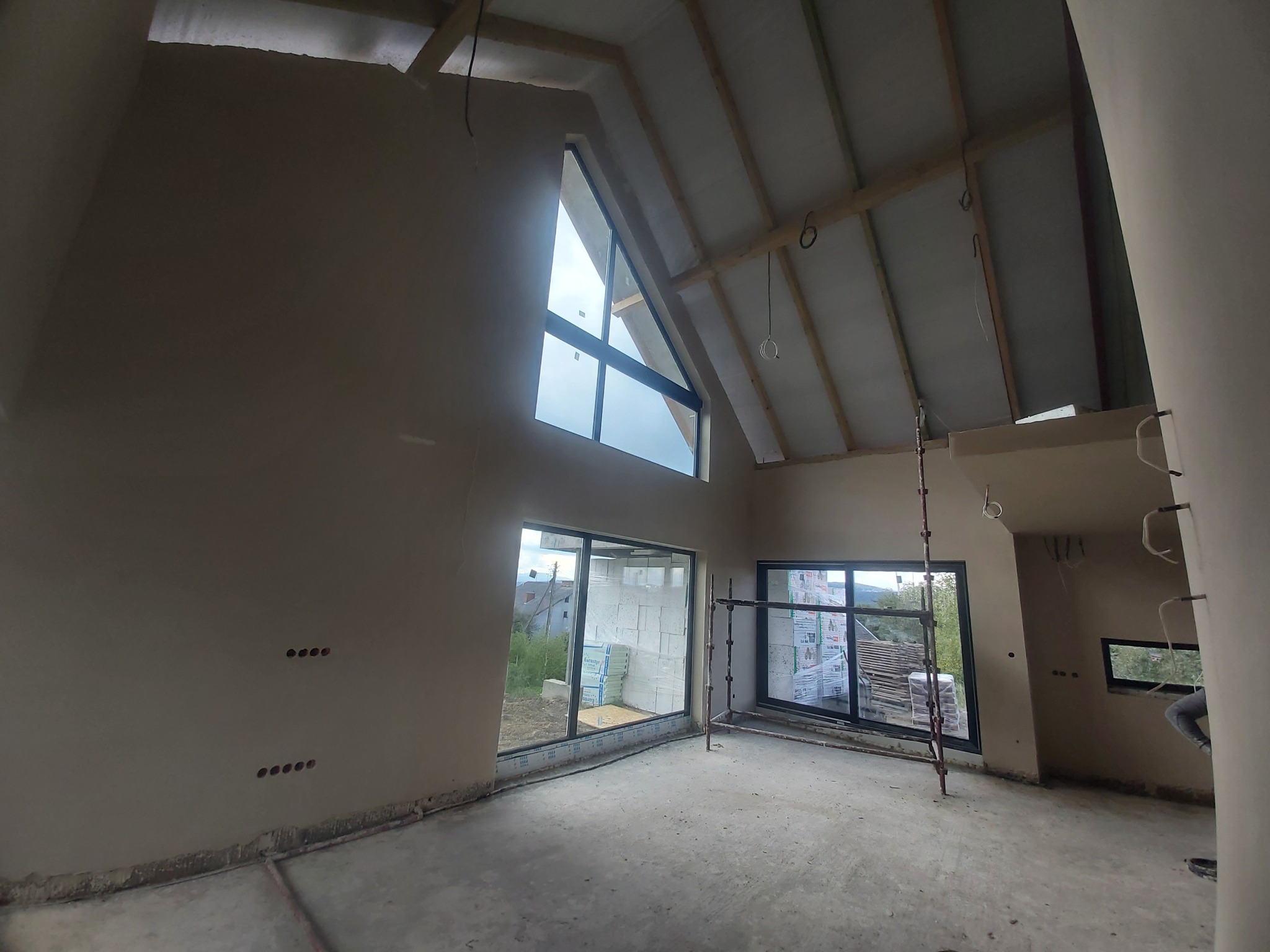 Wnętrze domu w trakcie prac wykończeniowych: ściany pokryte świeżym tynkiem, widoczne okna z widokiem na zewnątrz, rusztowanie w rogu pomieszczenia i przygotowane puszki elektryczne.